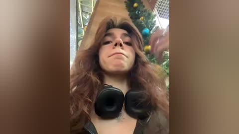 Media: A video of a young woman with long, wavy brown hair wearing black headphones, standing in a room adorned with a decorated Christmas tree. The background features a wooden staircase and festive decorations.