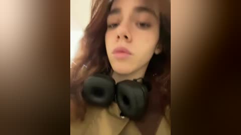 Media: A close-up video of a young person with fair skin, brown hair, and large black headphones, looking introspective. The background is blurry and brown-toned.