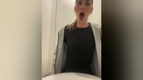 Media: Video of a Caucasian woman with light skin, brown hair, and hoop earrings, wearing a black top and open grey jacket, sticking her tongue out in a bathroom mirror.