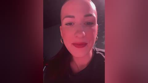 Media: Video of a bald, fair-skinned woman with red lipstick, wearing a black jacket, set against a dark, moody background.