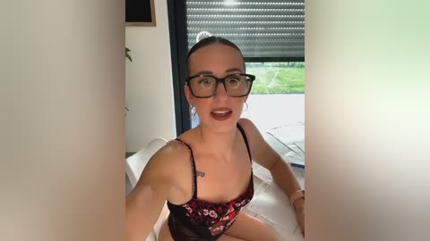 Media: A video of a fair-skinned woman with short brown hair, glasses, and a black floral bra, sitting on a bed with white sheets, taken from a selfie angle.