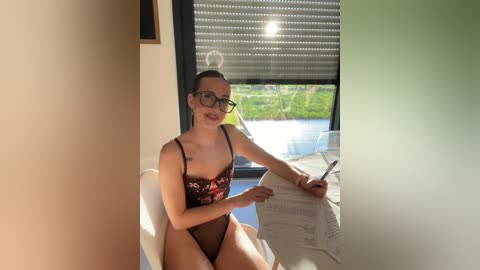 Media: Video of a slim, fair-skinned woman with glasses, wearing a black lace bra and panties, sitting on a white chair, writing on a white sheet of paper by a window with blinds partially open, revealing a green landscape outside.