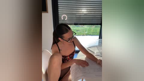 Media: Video of a woman with glasses and dark hair, wearing a red floral bra, sitting on a bed with a white blanket, reading a book. The background features a large window with blinds, revealing a green field outside.