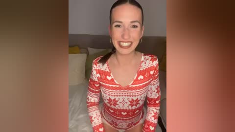 Media: Video of a smiling woman with light skin, brown hair in a ponytail, wearing a red and white Christmas-themed long-sleeve bodysuit, sitting on a bed with white sheets in a dimly lit room.