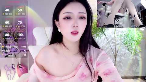 Media: A video featuring an East Asian woman with long black hair, wearing a pink floral off-shoulder top, sitting in a bright, modern room with white furniture. The background includes a green plant and a live stream overlay.