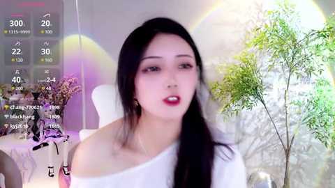 Media: A video of an East Asian woman with long black hair, wearing a white off-shoulder top, standing in a room with green plants and a white chair.