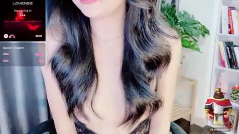 Media: Video of an Asian woman with long, wavy black hair, wearing a sheer, strapless top, partially exposing her breasts. Background features a bookshelf and potted plants.