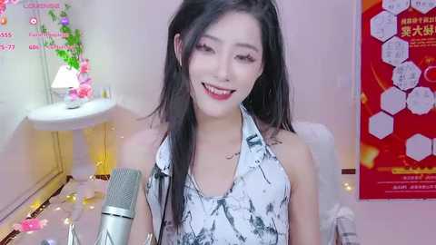 Media: A video of a smiling East Asian woman with long black hair, wearing a white halter top with a black abstract design, standing behind a microphone, surrounded by colorful decorations and a red poster in a cozy room.