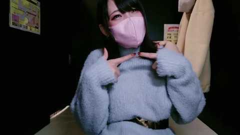 Media: Video of an Asian woman with long black hair, wearing a light blue fuzzy sweater, pink face mask, and a beige coat, indoors against a dark wall with a colorful chart.