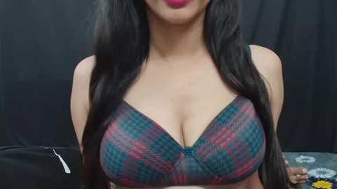 Media: Video of a woman with long, straight black hair, wearing a low-cut, patterned bra with pink and blue plaid. Her face is partially visible, lips painted a bold red. Black curtain background, blurred.