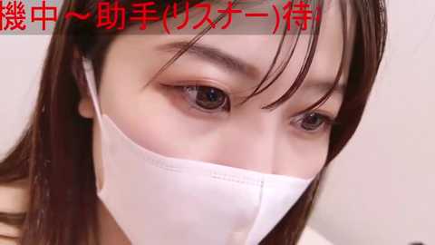 Media: Close-up video of a young Asian woman with straight, dark hair and brown eyes, wearing a white medical mask, with Japanese text overlay in red.