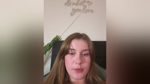 Video of a young woman with long, straight, light brown hair, fair skin, and a neutral expression. She is indoors, seated, with a potted plant and a decorative wall sign in the background.