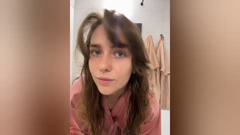 Media: Video of a young Caucasian woman with long, wavy brown hair and blue eyes, wearing a pink robe, standing in a modern bathroom with beige towels hanging on the wall.