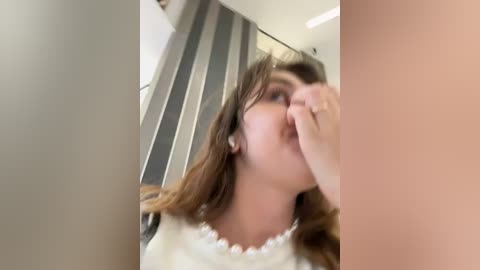 Media: Video of a young woman with light skin and long brown hair, wearing a white blouse, partially covering her face with her hand. The background features a modern, minimalist room with a gray and white striped curtain.