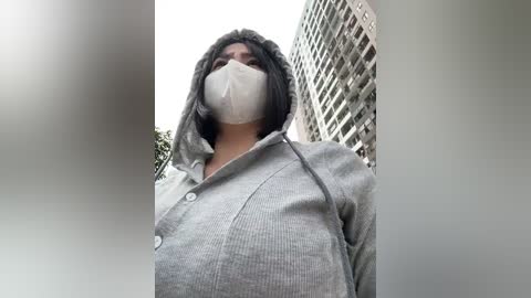 Media: Video of a person wearing a gray hoodie and white mask, standing in front of modern high-rise buildings with grid windows, blurred in the background.