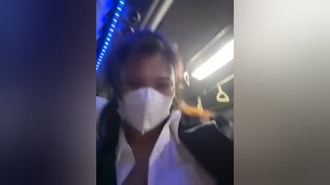 Media: Video of a blurred person wearing a white surgical mask, black jacket, and headphones, sitting in a dimly lit train with blue LED lights.