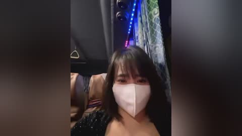 Media: A video of a young Asian woman with shoulder-length black hair, wearing a white face mask, lying on a bed, with a colorful patterned curtain and LED lights in the background.