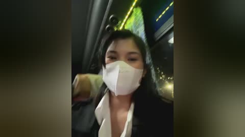 Media: Video of a woman wearing a white face mask and black jacket, holding a drink, in a dimly lit, modern interior.