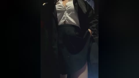 Media: A dimly lit video of a person in a black blazer over a white blouse, revealing a black skirt with a high slit. The person's face is not visible.