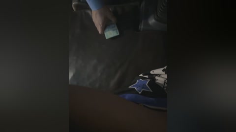 Media: A video showing a hand holding a smartphone, wearing a blue long-sleeved shirt, standing in a dimly lit room with a black, glossy surface. A blue and white star-patterned sneaker is visible in the foreground.