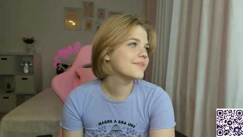 Video of a young Caucasian woman with shoulder-length blonde hair, light skin, and blue eyes, wearing a purple t-shirt, sitting in a pink gaming chair in a softly lit bedroom with a bed and white curtains.