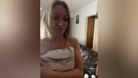 Media: Video of a fair-skinned, blonde woman, possibly in her late 20s, wrapped in a white towel, seated on a dark leather couch in a dimly lit room.