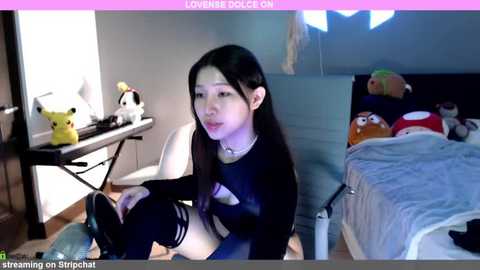 Media: Video of an Asian woman with long black hair in a black top and thigh-high socks, sitting on a chair in a cluttered bedroom with Pikachu plushies, a desk, and a bed.