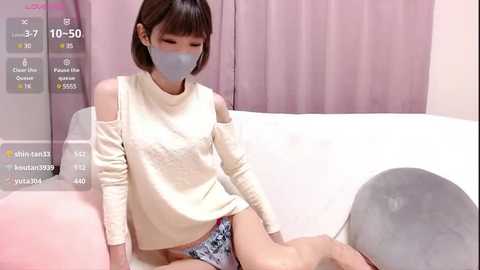 Media: Video of an Asian woman in a cream off-shoulder top, gray mask, and patterned panties, seated on a white sofa with a gray cushion. Background features light pink curtains and a digital screen displaying social media metrics.