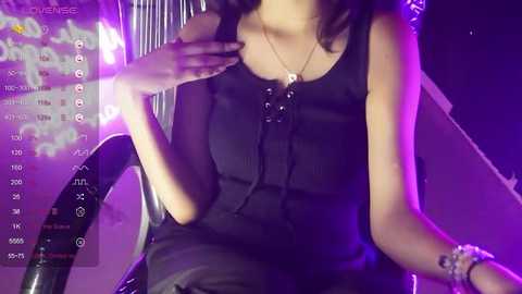 Media: A video of a woman with light skin and dark hair wearing a black sleeveless dress, sitting on a chair in a dimly lit room with purple lighting and a TV displaying a video feed.