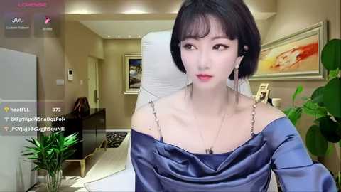 Video of a young Asian woman with short black hair, wearing an off-shoulder blue satin dress, sitting in a modern living room with beige walls, a white chair, and a potted plant.