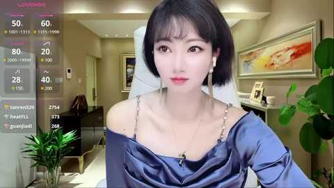 A video of a young East Asian woman with short black hair, wearing a blue satin off-shoulder dress, seated in a modern living room with a potted plant and abstract art on the wall.