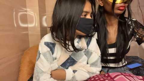 Media: Video of two Black women, one wearing a black face mask, white patterned sweater, and a denim jacket, the other with long black hair, black leather jacket, and a pink patterned shirt, seated indoors.