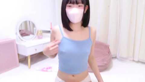 Media: Video of an East Asian woman with straight black hair and a pale complexion, wearing a blue crop top and white pants, standing in a pink-themed room with a vanity and white curtains.