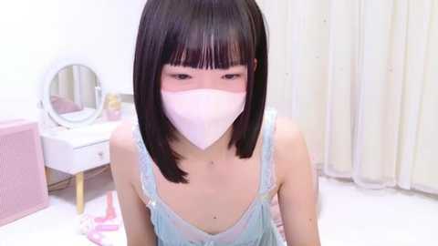 Media: Video of an East Asian woman with straight black hair, wearing a pink surgical mask, light blue spaghetti-strap dress, and sitting in a pastel-colored bedroom with a vanity mirror and white curtains.