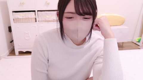 Media: Video of an Asian woman with short black hair, wearing a white face mask, a white sweater, and a white headband, sitting on a white bed in a bright, minimalist bedroom with white furniture and a yellow pillow.