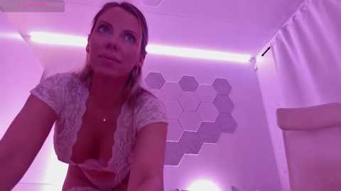 Media: Video of a blonde woman with fair skin, wearing a low-cut, lace-up top, in a room with purple lighting and hexagonal patterned wallpaper.