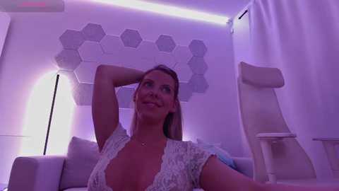 Media: Video of a smiling Caucasian woman with light skin, wearing a lace top, in a modern, purple-lit room with hexagonal wall art and white furniture.