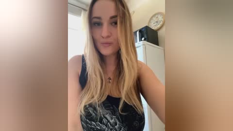 Media: Video of a blonde Caucasian woman with long hair, wearing a black floral-patterned tank top, making a pouty face. Background shows a white fridge, microwave, and clock in a kitchen.