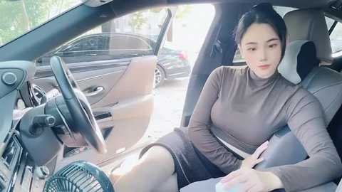 Media: Video of an Asian woman with long black hair in a brown turtleneck and black skirt, sitting in a car with the door open, looking outside at a parked silver sedan.