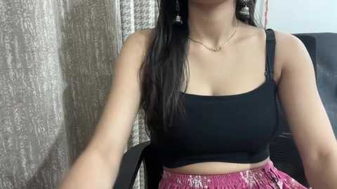 Media: Video of a young woman with long black hair, wearing a black crop top and pink patterned shorts, seated against textured beige curtains.