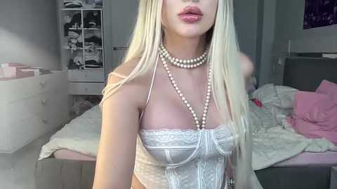 Media: Video of a slender, fair-skinned blonde woman in a white lace corset, pearl necklace, and pink lipstick, posing in a messy bedroom with a bed, dresser, and closet visible.