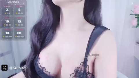 Media: Video of a fair-skinned woman with long black hair, wearing a black lace bra, showcasing her large breasts. The background is a blurred indoor setting with a calendar and a vase.