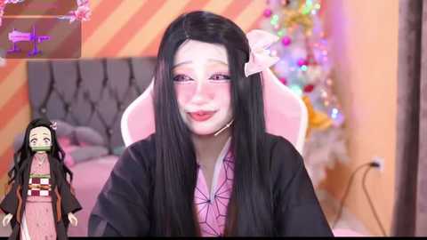 Media: Video of a Japanese anime character, Nezuko Kamado from \"Demon Slayer,\" with a soft, pastel pink and black cosplay outfit, seated in a pink chair. Background shows a festive, colorful room.