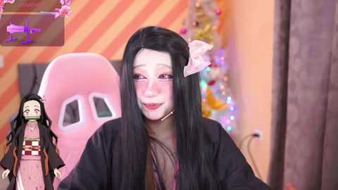 Media: Video of a woman wearing a black kimono with long black hair, sitting in a pink gaming chair. Background features a striped wall and a Christmas tree with colorful lights.