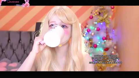 Media: Video of a young woman with long blonde hair, wearing a white mask, drinking from a white straw, in a festive room with a decorated Christmas tree.