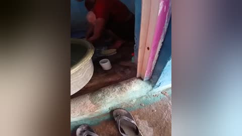 Media: Video of a small, dimly lit room with a person in a red shirt and dark pants kneeling on the floor, painting a wall. The room has a blue and green color scheme, and the floor is sandy. A white cup is nearby.