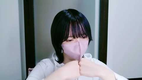 Media: Video of an Asian woman with straight black hair, wearing a pink face mask, and light blue shirt, praying with clasped hands in a modern, minimalist room with white walls.