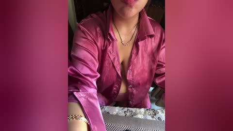 Media: Video of a woman with light skin, wearing a pink satin blouse with buttons partially unbuttoned, revealing cleavage, sitting on a floral-patterned cushion. She has a silver bracelet on her left wrist. Background is blurred.