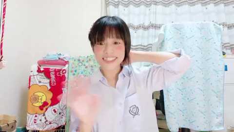 Media: A smiling young Asian woman with straight black hair and bangs, wearing a light purple button-up shirt, poses in a brightly lit room with floral curtains and patterned bedding.