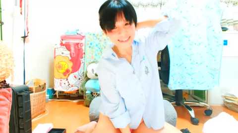 Media: Video of a cheerful young woman with short black hair, wearing a light blue shirt, holding a blue towel in a brightly lit, cluttered bedroom with a wicker basket and laundry.